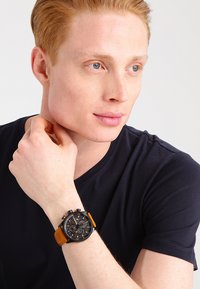 Orologio cronografo nero con cinturino in pelle arancione, quadrante rotondo e diversi quadranti più piccoli; indossato su un polso con una maglietta blu navy.