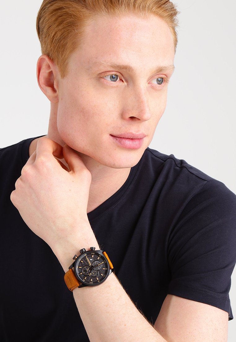 Orologio cronografo nero con cinturino in pelle arancione, quadrante rotondo e diversi quadranti più piccoli; indossato su un polso con una maglietta blu navy.