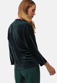Dunkelgrünes Samt-Oberteil mit Dreiviertelärmeln und lockerer Passform. Verfügt über eine glatte Textur und dezente Drapierung auf der Rückseite.