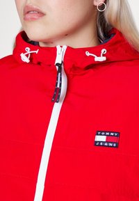 Czerwona kurtka z nylonu ma białe zapięcie na zamek, regulowany kaptur z białymi uszami oraz niebiesko-białe haftowane logo "TOMMY JEANS" na piersi.