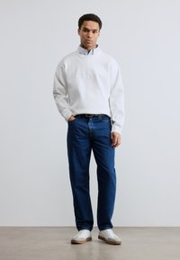 Άσπρη φαρδιά μπλούζα με ριμπ λαιμόκοψη, μπλε τζιν και άσπρα αθλητικά παπούτσια με μπεζ σόλες· άνδρας ποζάρει όρθιος με τα χέρια στις τσέπες.