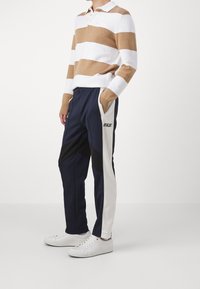 Randig stickad polo i vitt och beige, kombinerad med navyblå och krämfärgade träningsbyxor med svarta accenter och logo. Vita sneakers fulländar outfiten.