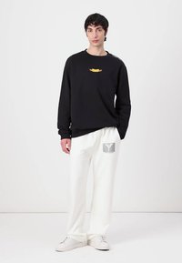Sort sweatshirt med lange ærmer og gul grafik samt hvide manchet-detaljer, parret med hvide sweatpants med en grå logo-patch. Hvide sneakers.