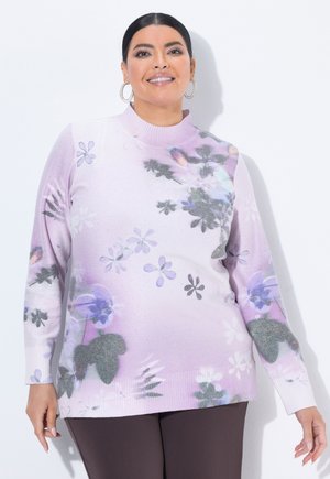 Smilende kvinde med øreringe formet som ringe iført en pastel lilla blomsterstrikket sweater og mørke bukser mod en hvid baggrund.