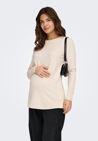 Έγκυος γυναίκα με κυματιστά καστανά μαλλιά φοράει μπεζ μπλούζα μακρυμάνικη με κουμπιά στο πλάι, μαύρο παντελόνι και μαύρη τσάντα ώμου.