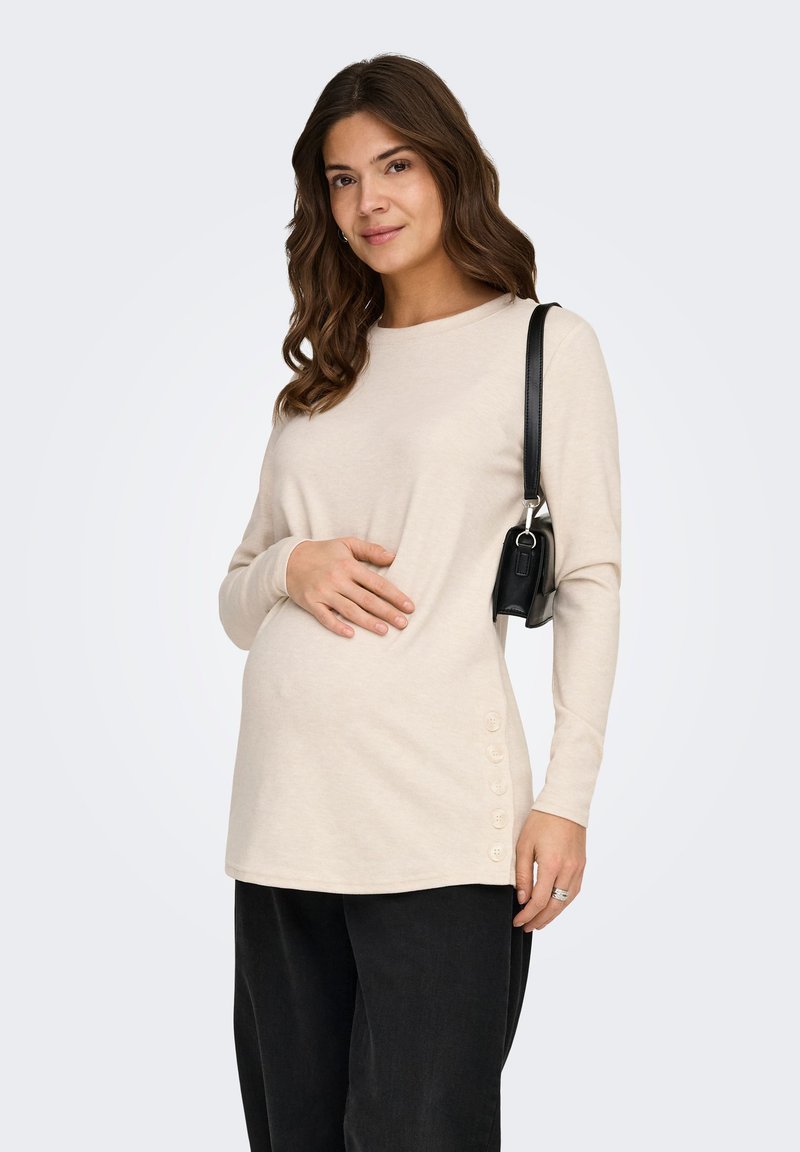 Έγκυος γυναίκα με κυματιστά καστανά μαλλιά φοράει μπεζ μπλούζα μακρυμάνικη με κουμπιά στο πλάι, μαύρο παντελόνι και μαύρη τσάντα ώμου.