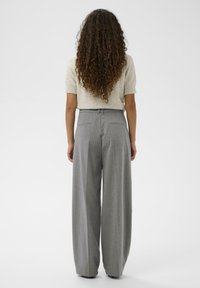 Kobieta z długimi kręconymi włosami ma na sobie beżowy top z krótkim rękawem oraz wysokie, szerokie spodni w szaro-białe paski, stoi na tle białej ściany.