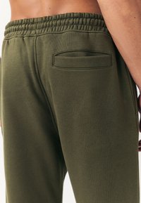 Alyvuogių žali spalvos sportinės kelnės su tekstūruota paviršiaus struktūra, elastiniu juosmeniu ir maža užpakaline kišene, pasižyminčios prigludusiu dizainu ir lygiu audiniu.