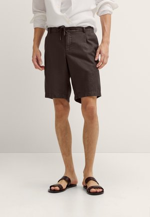 Mann trägt dunkelbraune knielange Shorts, weißes hochgekrempeltes Langarmhemd und dunkelbraune Sandalen und steht vor einem schlichten Hintergrund.