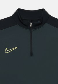 Nike pulover u tamnozelenoj tkanini s visokim okovratnikom, zatvaranjem na pola munje, crnim naglascima na ramenima i žutim logotipom na lijevoj strani prsnog koša.