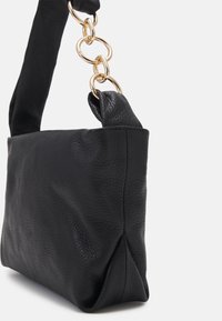Sac à main en cuir noir texturé avec une sangle douce et rembourrée attachée par de larges maillons de chaîne dorés sur un fond clair uni.