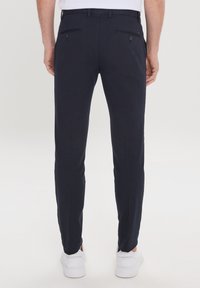 Pantalon de marine ajusté, fabriqué en tissu lisse. Dispose de deux poches arrière avec boutons et d'une taille plate. Chaussures visibles en bas.