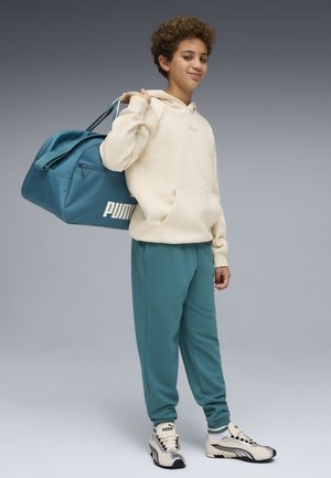 Ung person iført en beige hættetrøje og blågrønne bukser, bærende en blågrøn Puma rejsetaske over skulderen, stående foran en grå baggrund.