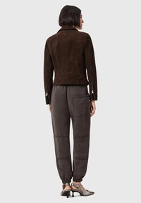 Giacca in suede marrone con vestibilità su misura e polsini con zip, abbinata a pantaloni grigi larghi con cintura elastica e tasche laterali.