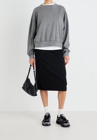 Persona vistiendo un sudadera gris lavada, una falda negra hasta las rodillas, calcetines blancos, zapatillas negras y sosteniendo un bolso negro frente a un fondo blanco.