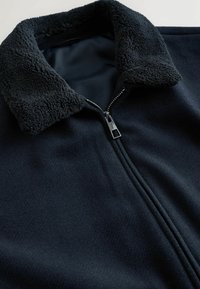 Musta fleece-takki, jossa on teksturoitu kaulus ja hopeinen vetoketju, kuvattu osittain vetoketjusta ja tasaisena valkoista taustaa vasten.