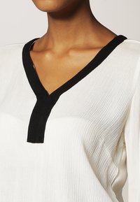 Haut blanc avec une finition texturée, présentant un col en V noir et une patte partielle. Le tissu est léger et fluide.