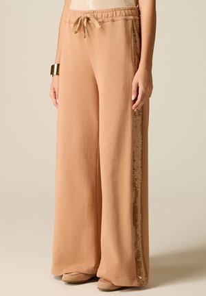 Beżowe spodnie o szerokich nogawkach z elastycznym pasem i sznurkiem, z cekinowym paskiem z boku, noszone z beżowymi mokasynami i złotą bransoletą.