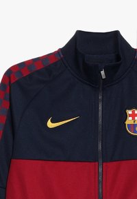 Nike vetoketjullinen takki, jossa on tummansinisiä ja punaisia paneeleja, ruudullinen kuvio harteilla, kultainen logo ja seuran vaakuna rinnassa.