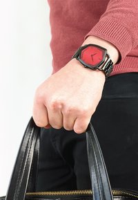 Rød hexagonal ur med et sort metallisk bånd. Urskiven har minimalistiske timemarkører og visere. Sort læderhandbag holdt i den anden hånd.