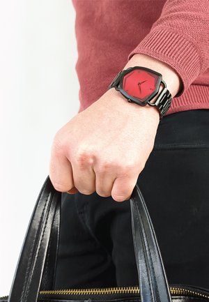 Montre hexagonale rouge avec un bracelet métallique noir. Le cadran de la montre présente des marqueurs d'heure et des aiguilles minimalistes. Sac à main en cuir noir tenu dans l'autre main.