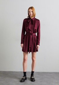 Vestido de satén burdeos con mangas largas, detalles fruncidos en el frente y un cinturón en la cintura. Combinado con calcetas negras y mocasines.