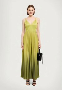 Femme portant une robe maxi sans manches, plissée, en dégradé de vert, tenant un petit sac à main noir, se tenant devant un fond uni.