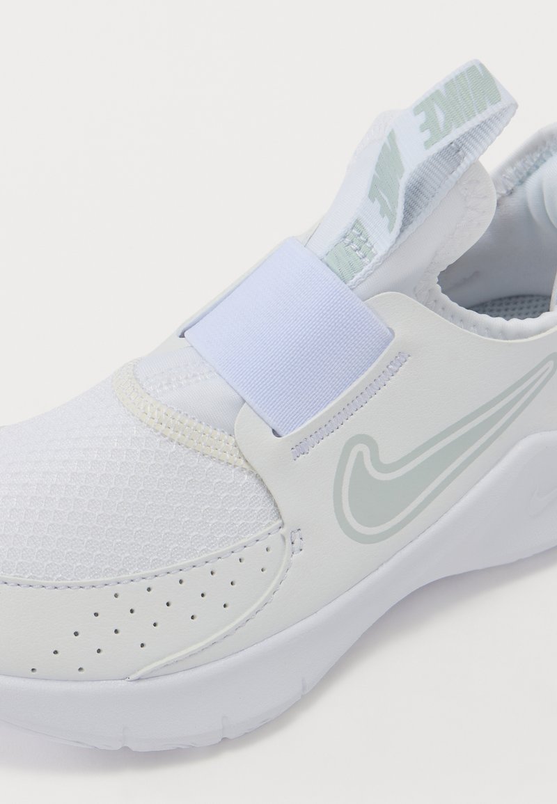 Zapatilla deportiva blanca con parte superior de malla, correa elástica y puntera redonda. Presenta un logo discreto y perforaciones para transpirabilidad.