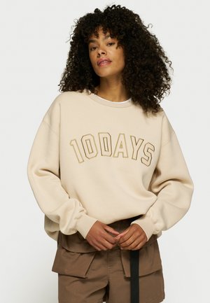 Sudadera beige con la inscripción "10DAYS" en letras negritas. Presenta un corte holgado, mangas oversize y una tela suave y texturizada. Combinada con pantalones cortos marrones.