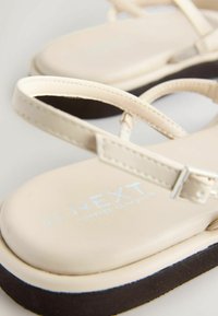 Bézs lapos szandálok vékony pántokkal és kis csattal, párnázott talpbetéttel és fekete talppal.