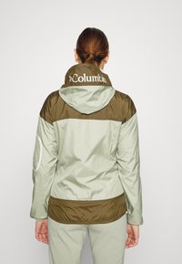 Mujer con cabello castaño recogido en un moño, vistiendo una chaqueta Columbia bicolor verde con capucha, de espaldas mostrando la parte trasera de la chaqueta y el nombre de la marca.