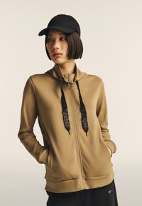 Sudadera con cremallera color caqui, con cordones negros, mangas establecidas y bolsillos tipo canguro, combinada con una gorra de béisbol negra y pantalones oscuros.