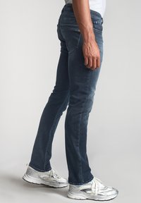Jeans slim-fit bleu foncé en denim, avec une texture délavée et un design à cinq poches. Portés avec des baskets métalliques argentées.