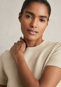 Maglione beige lavorato a maglia con maniche corte e scollatura a costine. La mano del modello è appoggiata sulla spalla, mostrando un tono di pelle uniforme e un trucco naturale.
