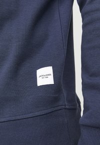 Felpa blu navy con orlo a coste, dotata di un'etichetta in tessuto bianco con testo nero che recita "JACK&JONES EST. 1990." Texture liscia.