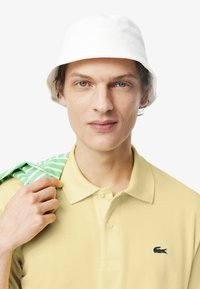 Polo giallo con colletto, dotato di un piccolo logo ricamato verde. Un cappello a secchiello bianco e una camicia a strisce verdi appoggiata sulla spalla.
