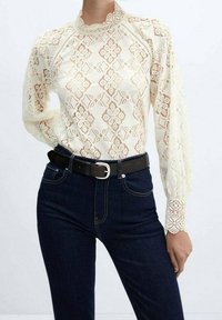Spidst blouse i creme med geometrisk mønster og høj halsudskæring. Lange ærmer med spidskant. Kombineret med mørke jeans og et sort bælte.