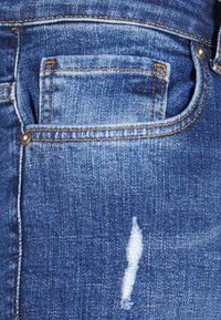 Jeans iz denima v srednjem modrem odtenku, z majhnim sprednjim žepom, vidnim šivom v oranžni barvi in unikatnim podrobnostmi z belim nitom.