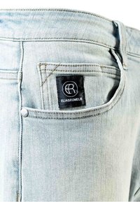 Jean en denim bleu clair avec une poche arrière et une étiquette noire portant le nom de la marque « ELIASRUMELIS », bords cousus et détail de bouton argenté.