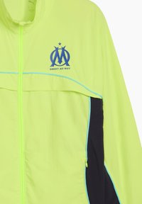 Neon gul vindjacka med blå accenter, med dragkedja fram, logotyp och en mörkblå sidopanel. Lätt och strukturerad tyg.