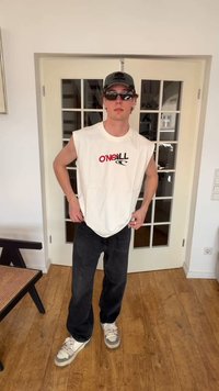 Jeune homme portant une casquette noire, des lunettes de soleil, un débardeur blanc O'NEILL, un pantalon noir et des baskets, se tenant à l'intérieur devant des portes doubles vitrées.