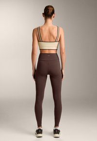 Beigen värinen rintaliivitoppi, jossa tummat olkaimet, yhdistetty korkeavyötäröisiin ruskeisiin leggingsseihin. Sileä pinta, vartalonmyötäinen malliltaan ja nilkkaan ulottuva pituus.