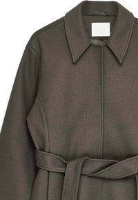 Tumši zaļa virsjaka ar strukturētu apkakli, jostu viduklī un gludu, teksturētu audumu. Priekšpuses aizdare un minimālistisks dizains.