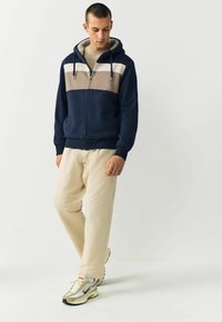 Námornícky zip-up mikina s béžovými a bielymi horizontálnymi pruhmi, mäkké podšívku, spárovaná so smotanovými nohavicami a béžovými teniskami.