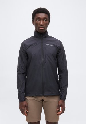 Homme portant une veste noire zippée avec une poche poitrine et un short beige, debout devant un fond blanc uni.