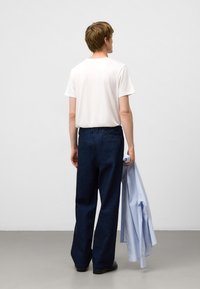 Junger Mann steht mit dem Rücken zur Kamera, trägt ein weißes T-Shirt und dunkelblaue Jeans und hält ein hellblaues Hemd in seiner rechten Hand.