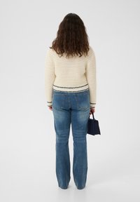 Stickad cremefärgad tröja med ett strukturerat mönster och svart kant, kombinerad med ljusblå utsvängda jeans. En liten mörk handväska hålls i ena handen.