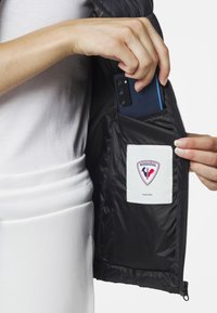 Innenansicht einer schwarzen gepolsterten Jacke mit einem weißen Stoffetikett, das ein Logo zeigt. Ein blauer Smartphone wird in eine Seitentasche gelegt.