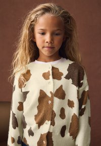 Pleteni cardigan v kremni barvi z rjavim vzorcem kravjih pik, z gumbi spredaj in rebrastimi manšetami. Mehka tekstura in sproščen kroj.