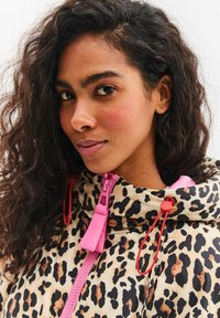 Bunda s leopardím potiskem na béžovém pozadí, růžovým zipem a červenými detaily. Má vysoký límec a stahovací šňůrku pro nastavitelnost.